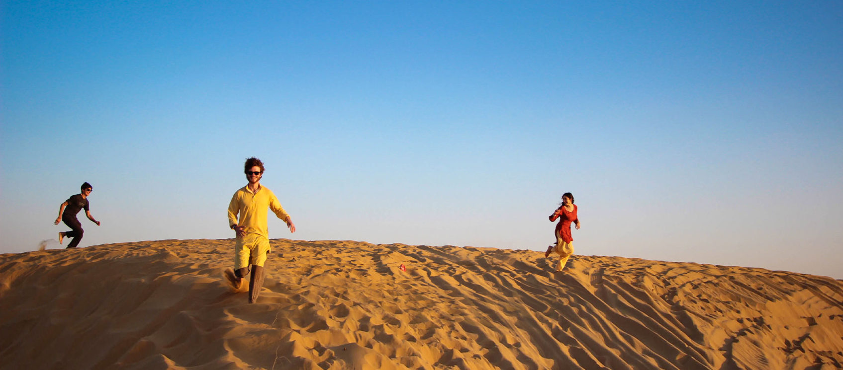 Man in yellow and woman in red running through sand dunes (day)