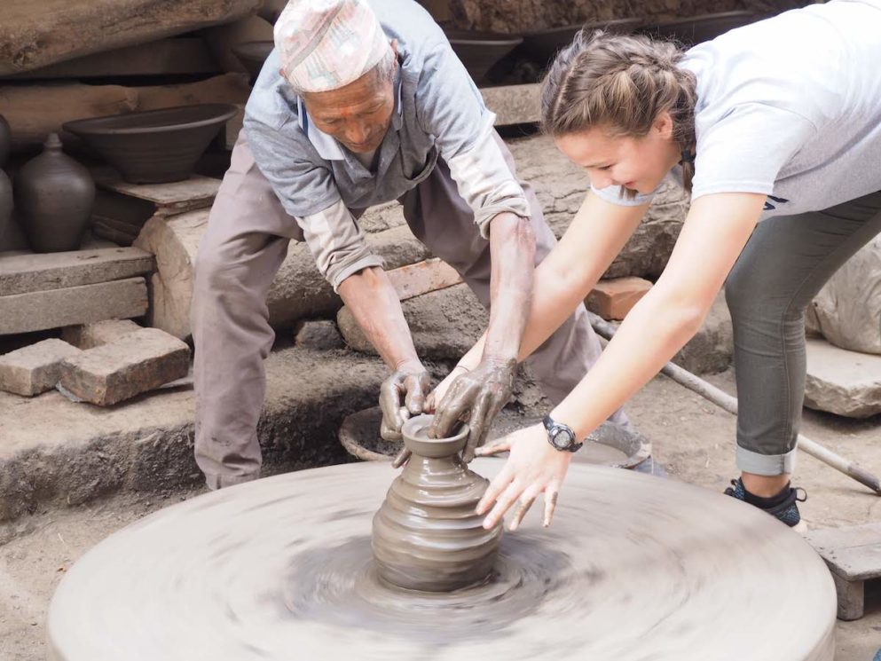 man in nepal teaching young white woman to do pottery on a wheel